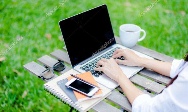 Freelance work. Casual dressed man sitting at wooden desk inside garden working on computer pointing with color pen electronic gadgets dropped around on table side view